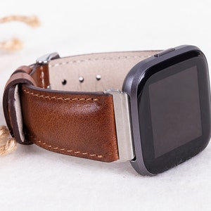 May include: A brown leather watch band with silver hardware attached to a black smartwatch with a square face.
