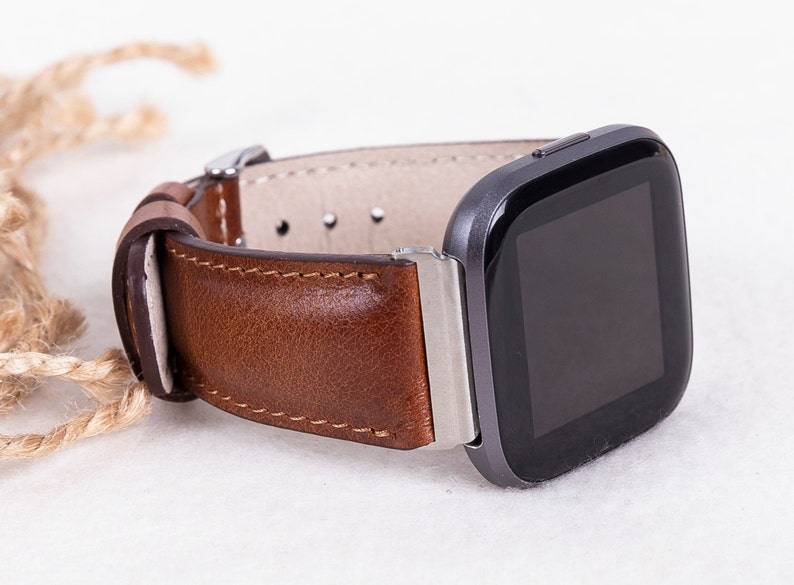 May include: A brown leather watch band with silver hardware attached to a black smartwatch with a square face.