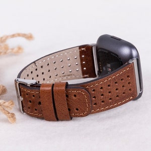 May include: A brown leather watch band with a perforated design. The band is attached to a silver watch buckle and a gray smartwatch.