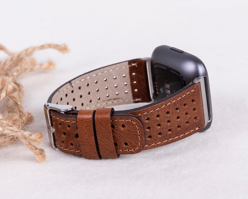 May include: A brown leather watch band with a perforated design. The band is attached to a silver watch buckle and a gray smartwatch.
