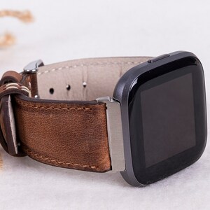 Puede incluir: Una correa de reloj de cuero marr&oacute;n con una hebilla plateada y un reloj inteligente cuadrado negro con una pantalla negra.