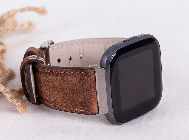 Puede incluir: Una correa de reloj de cuero marr&oacute;n con una hebilla plateada y un reloj inteligente cuadrado negro con una pantalla negra.