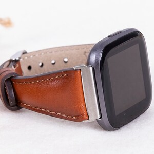 May include: A brown leather watch strap with a silver buckle attached to a black smartwatch with a square face.