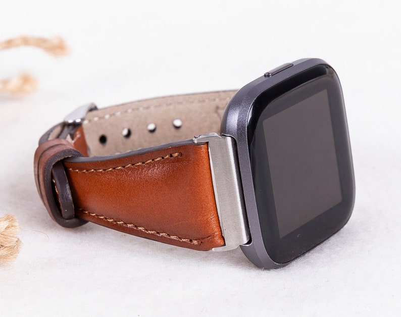 May include: A brown leather watch strap with a silver buckle attached to a black smartwatch with a square face.