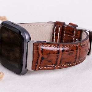 May include: A brown leather watch band with a crocodile pattern. The band is designed for a square-shaped smartwatch.