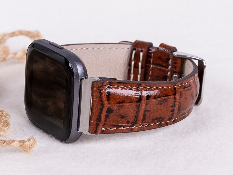 May include: A brown leather watch band with a crocodile pattern. The band is designed for a square-shaped smartwatch.