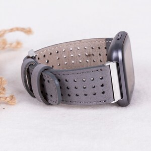 Puede incluir: Correa de reloj de cuero gris con un dise&ntilde;o perforado. La correa se muestra unida a un reloj inteligente.