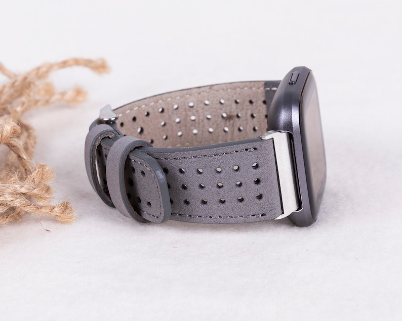 Puede incluir: Correa de reloj de cuero gris con un dise&ntilde;o perforado. La correa se muestra unida a un reloj inteligente.