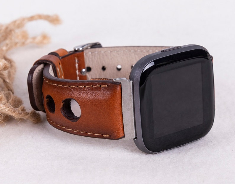 May include: A brown leather watch band with stitching and holes for a smartwatch. The band is attached to a black smartwatch with a square face.