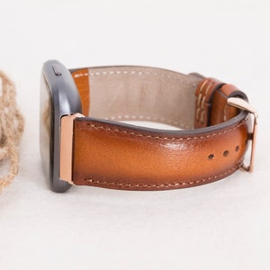 Op de afbeelding: Een bruin leren horlogebandje met ros&eacute;gouden hardware. Het bandje is bevestigd aan een smartwatch.