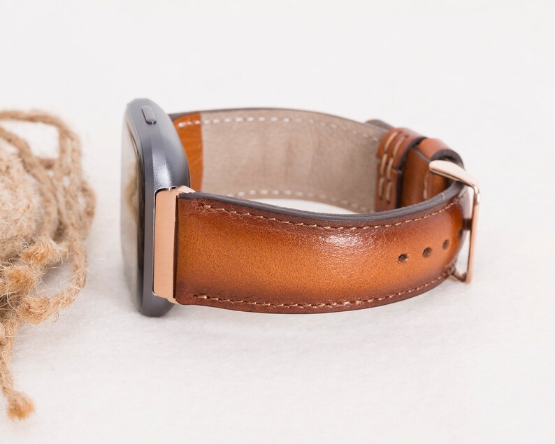 Op de afbeelding: Een bruin leren horlogebandje met ros&eacute;gouden hardware. Het bandje is bevestigd aan een smartwatch.
