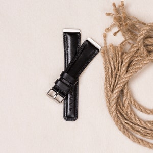 May include: Two black leather watch straps with silver-colored metal clasps and end pieces are displayed on a cream-colored surface. A length of coiled, natural-colored rope is next to the straps.
