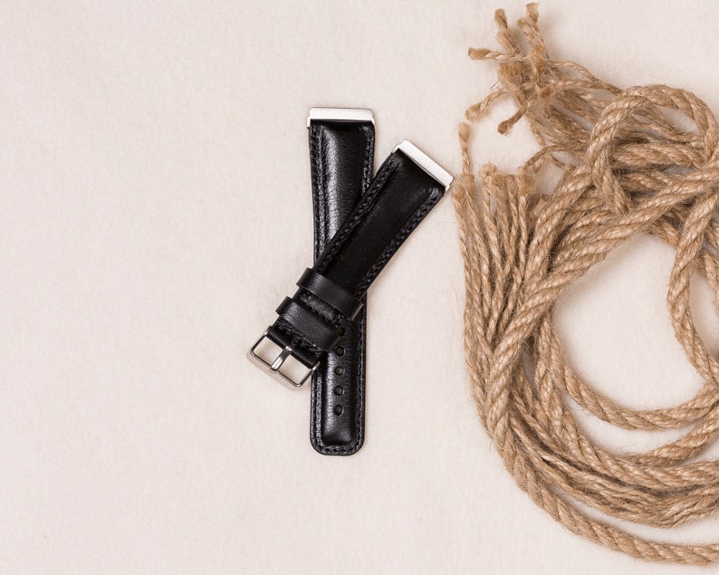 May include: Two black leather watch straps with silver-colored metal clasps and end pieces are displayed on a cream-colored surface. A length of coiled, natural-colored rope is next to the straps.