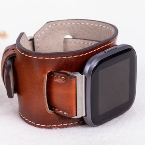 K&ouml;nnte beinhalten: Ein braunes Lederarmband mit einer silbernen Schnalle und einer schwarzen Smartwatch.