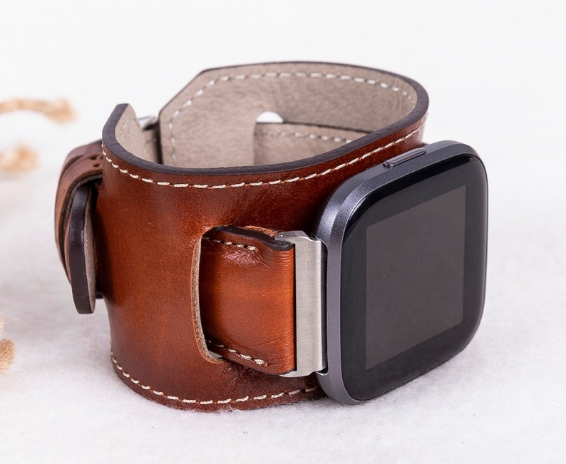 K&ouml;nnte beinhalten: Ein braunes Lederarmband mit einer silbernen Schnalle und einer schwarzen Smartwatch.