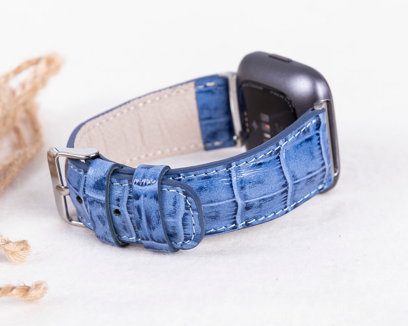 Puede incluir: Una correa de reloj de cuero azul claro con un patr&oacute;n de cocodrilo. La correa est&aacute; dise&ntilde;ada para un reloj inteligente con una cara cuadrada.