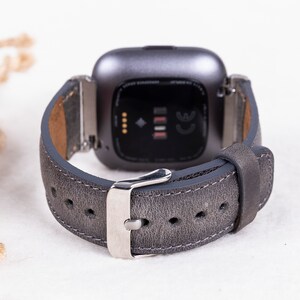 May include: A gray leather watch band with a silver buckle. The band is designed for a smartwatch with a square face.