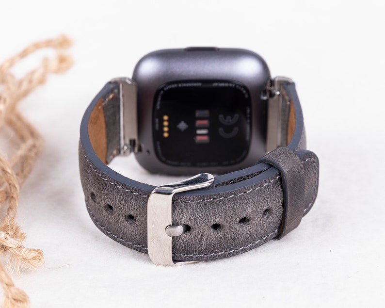 May include: A gray leather watch band with a silver buckle. The band is designed for a smartwatch with a square face.