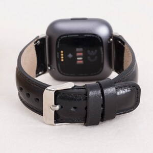 May include: A black leather watch band with a silver buckle is attached to a gray smartwatch. The watch band is made of smooth, dark leather. The watch face is dark gray with a black screen.
