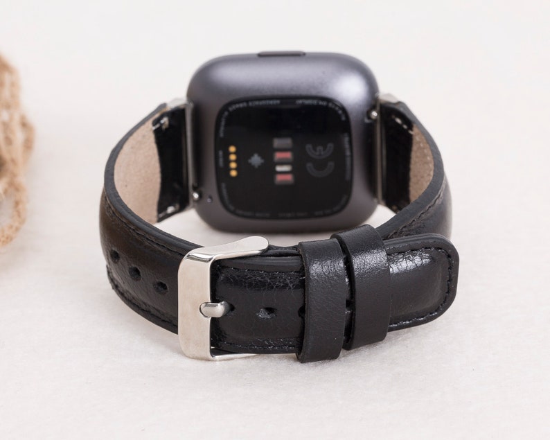 May include: A black leather watch band with a silver buckle is attached to a gray smartwatch. The watch band is made of smooth, dark leather. The watch face is dark gray with a black screen.