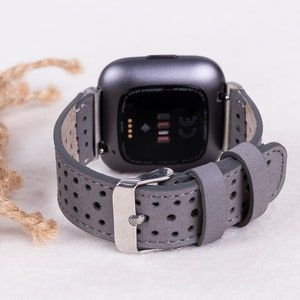 Puede incluir: Correa de reloj de cuero gris con un dise&ntilde;o perforado y una hebilla plateada. La correa es compatible con los relojes inteligentes Fitbit Versa.