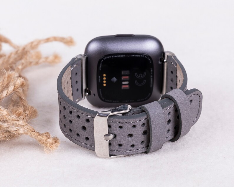 Puede incluir: Correa de reloj de cuero gris con un dise&ntilde;o perforado y una hebilla plateada. La correa es compatible con los relojes inteligentes Fitbit Versa.