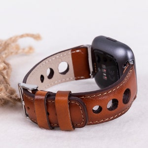 May include: A brown leather watch band with a silver buckle and a gray fitness tracker. The band has a perforated design and is designed for a smart watch.