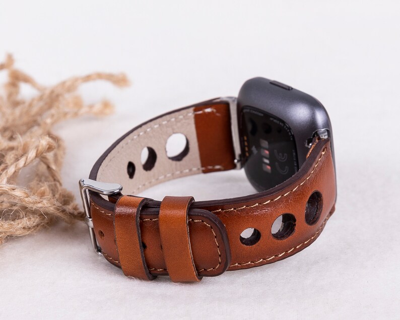 May include: A brown leather watch band with a silver buckle and a gray fitness tracker. The band has a perforated design and is designed for a smart watch.