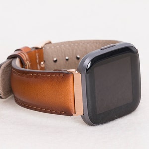 Op de afbeelding: Een bruin leren horlogebandje met een ros&eacute;gouden gesp en een zwart vierkant smartwatch.