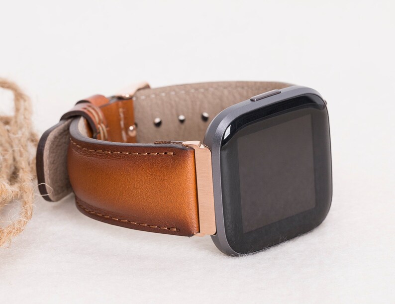 Op de afbeelding: Een bruin leren horlogebandje met een ros&eacute;gouden gesp en een zwart vierkant smartwatch.