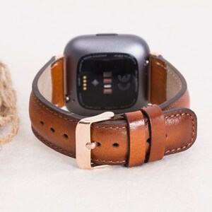 Op de afbeelding: Een bruin leren horlogebandje met een ros&eacute;gouden gesp. Het bandje is ontworpen voor een smartwatch met een vierkant gezicht.