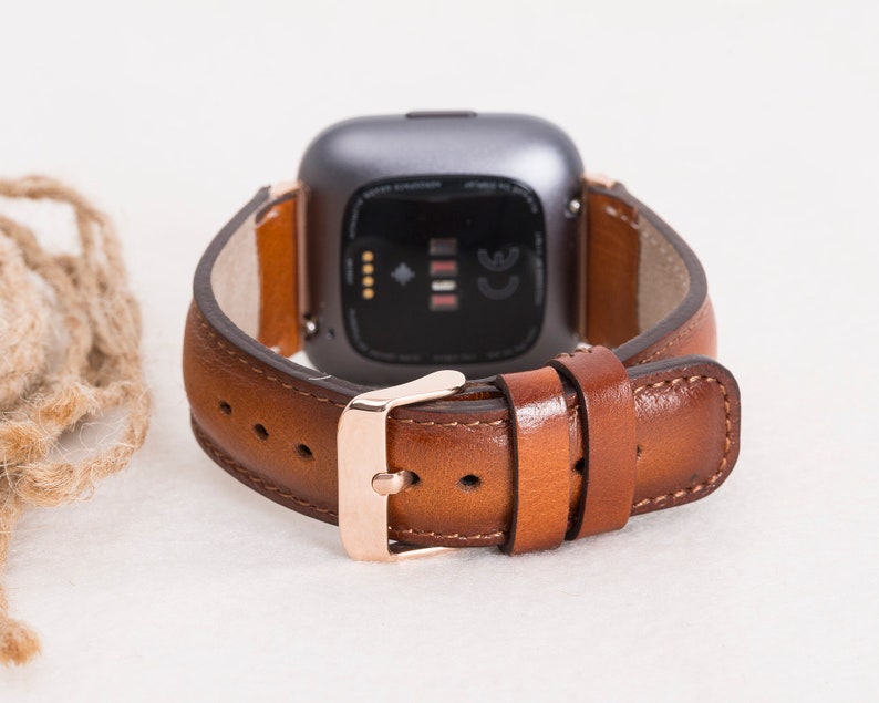 Op de afbeelding: Een bruin leren horlogebandje met een ros&eacute;gouden gesp. Het bandje is ontworpen voor een smartwatch met een vierkant gezicht.
