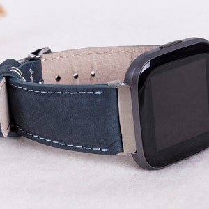 May include: A dark blue leather watch band with white stitching, attached to a black smartwatch with a square face.