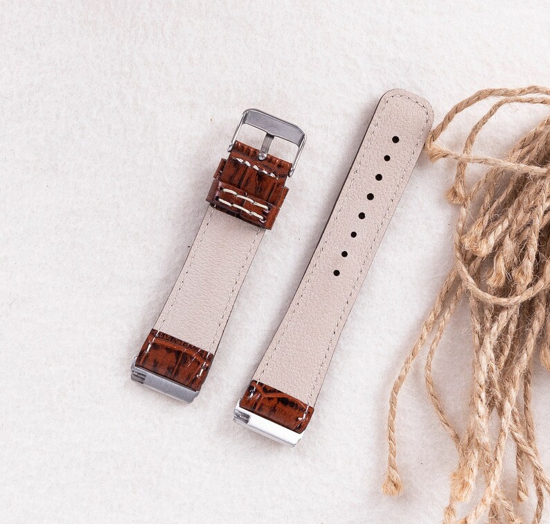 May include: Two brown leather watch straps with silver buckles. The straps have a crocodile pattern and are laid out on a white surface.
