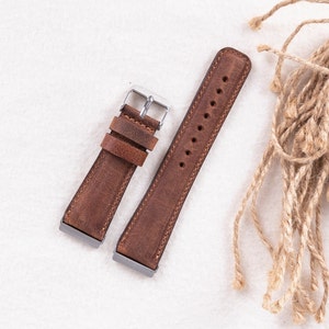 Puede incluir: Dos correas de reloj de cuero marr&oacute;n con hebillas plateadas. Las correas est&aacute;n dispuestas sobre una superficie blanca.