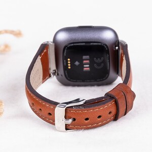 May include: A brown leather watch strap with a silver buckle and a silver smartwatch with a black screen and the CE mark.