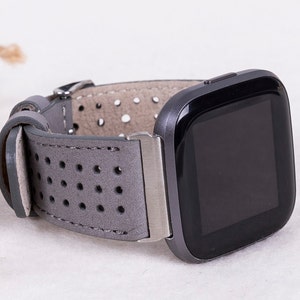 Puede incluir: Una correa de reloj de cuero gris con un dise&ntilde;o perforado. La correa est&aacute; unida a un reloj inteligente negro con una esfera cuadrada.