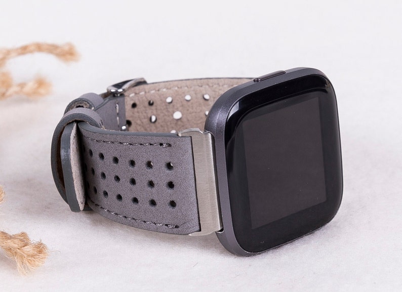 Puede incluir: Una correa de reloj de cuero gris con un dise&ntilde;o perforado. La correa est&aacute; unida a un reloj inteligente negro con una esfera cuadrada.