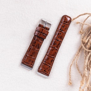 May include: Two brown leather watch straps with a crocodile pattern. The straps have silver buckles and are laid out on a white surface.