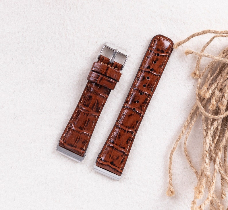 May include: Two brown leather watch straps with a crocodile pattern. The straps have silver buckles and are laid out on a white surface.