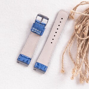 Puede incluir: Dos correas de reloj con cuero azul claro y costuras blancas. Las correas son de material beige claro y tienen una hebilla plateada.