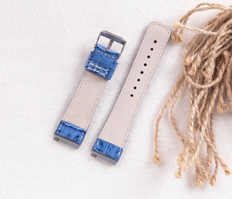 Puede incluir: Dos correas de reloj con cuero azul claro y costuras blancas. Las correas son de material beige claro y tienen una hebilla plateada.
