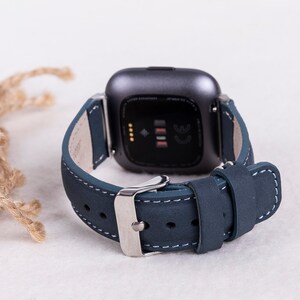 May include: A dark blue leather watch band with white stitching and a silver buckle. The band is designed for a square-shaped smartwatch.