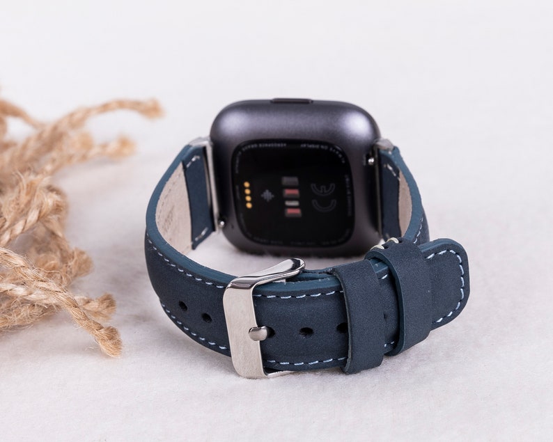 May include: A dark blue leather watch band with white stitching and a silver buckle. The band is designed for a square-shaped smartwatch.