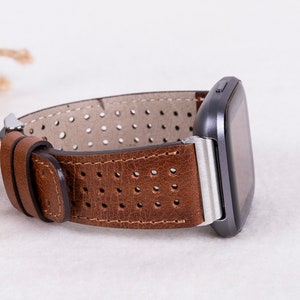 May include: Brown leather watch band with a perforated design and a silver buckle. The band is attached to a black smartwatch.