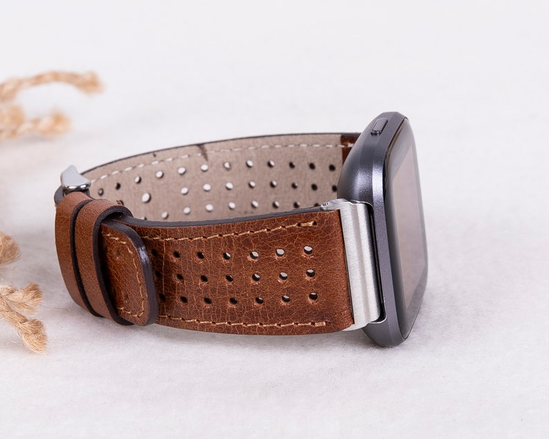 May include: Brown leather watch band with a perforated design and a silver buckle. The band is attached to a black smartwatch.