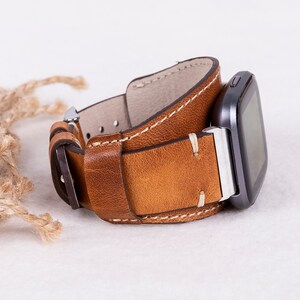 May include: A brown leather watch strap with white stitching. The strap is attached to a black smartwatch with a silver buckle.