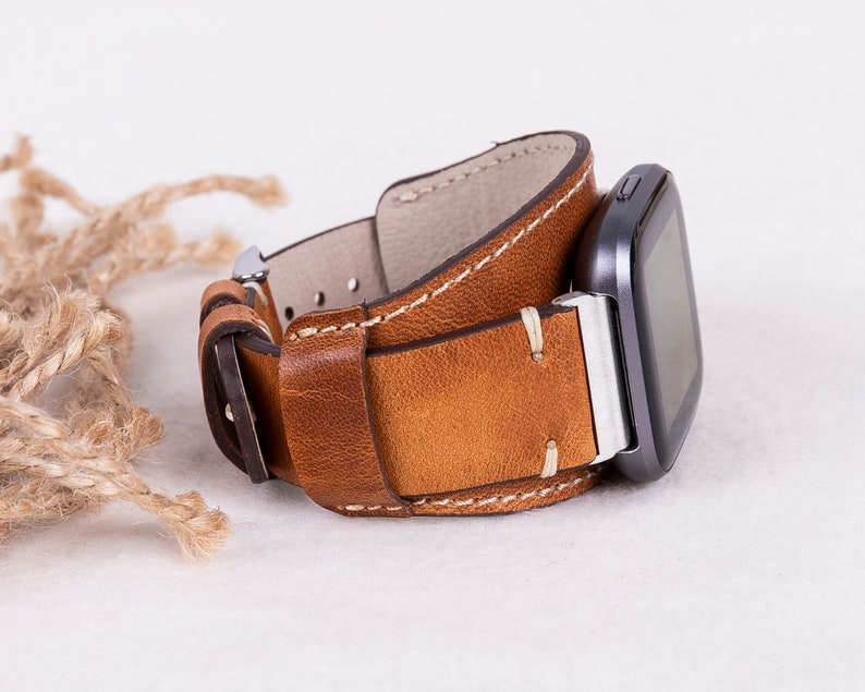 May include: A brown leather watch strap with white stitching. The strap is attached to a black smartwatch with a silver buckle.