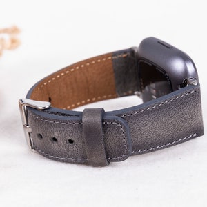 May include: A gray leather watch band with a silver buckle. The band is designed for a smartwatch with a square face.