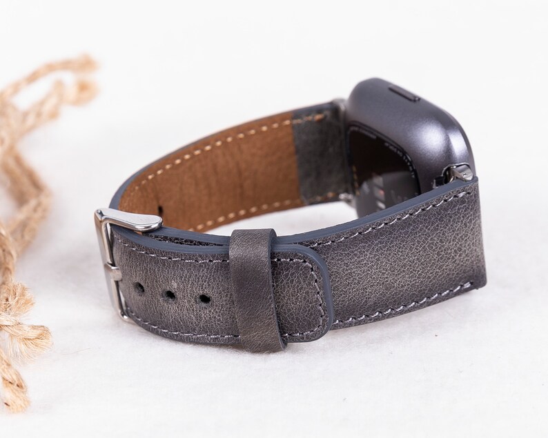 May include: A gray leather watch band with a silver buckle. The band is designed for a smartwatch with a square face.